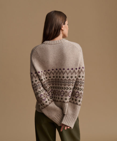 A woman shown from the back wearing a beige knit sweater with a dark brown and purple Fair Isle pattern across the shoulders and sleeves, paired with olive green pants, against a solid beige studio background.