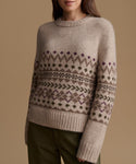 A woman wearing a beige knit sweater with a brown and purple Fair Isle pattern, paired with olive green pants, against a solid beige background.