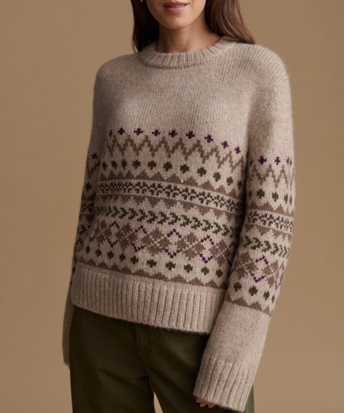A woman wearing a beige knit sweater with a brown and purple Fair Isle pattern, paired with olive green pants, against a solid beige background.