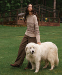 A woman stands on a grassy lawn wearing a beige knit sweater with a brown geometric pattern and brown wide-leg pants, while standing next to a large fluffy white dog. In the background, there is a wooden greenhouse structure and trees.