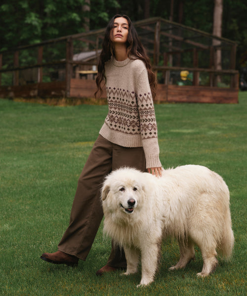 A woman stands on a grassy lawn wearing a beige knit sweater with a brown geometric pattern and brown wide-leg pants, while standing next to a large fluffy white dog. In the background, there is a wooden greenhouse structure and trees.