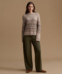 A woman stands against a plain beige studio background wearing a light taupe knit sweater with a dark brown Fair Isle pattern across the chest and sleeves, paired with olive green wide-leg trousers and brown suede shoes.