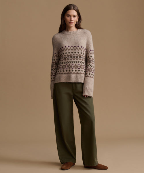 A woman stands against a plain beige studio background wearing a light taupe knit sweater with a dark brown Fair Isle pattern across the chest and sleeves, paired with olive green wide-leg trousers and brown suede shoes.