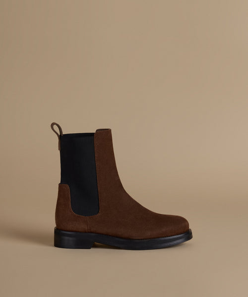 A brown suede Chelsea boot with black elastic side panels and a black sole, shown in profile against a solid beige background.