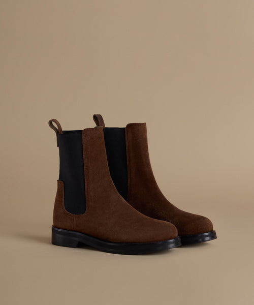A pair of brown suede Chelsea boots with black elastic side panels and black soles, displayed on a warm beige background.