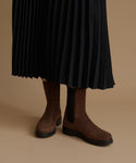 A person wearing dark brown suede Chelsea boots with black elastic side panels, paired with a black pleated midi skirt, against a solid beige studio background.