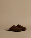 A pair of dark brown slip-on mule clogs with a soft cream sherpa lining, displayed on a warm beige studio background.