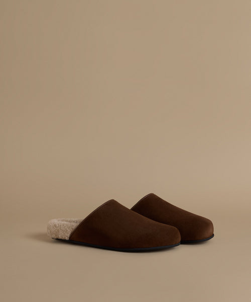 A pair of dark brown slip-on mule clogs with a soft cream sherpa lining, displayed on a warm beige studio background.