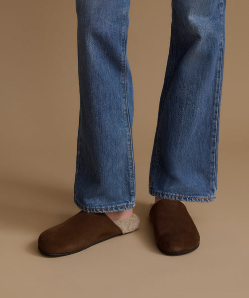 A person wearing blue jeans and dark brown suede slip-on slippers with a beige knit lining, standing against a solid tan background.