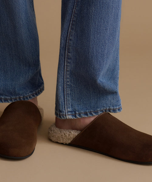 A close-up of a person wearing brown suede mule slippers with beige sherpa lining, paired with blue jeans, against a solid beige background.