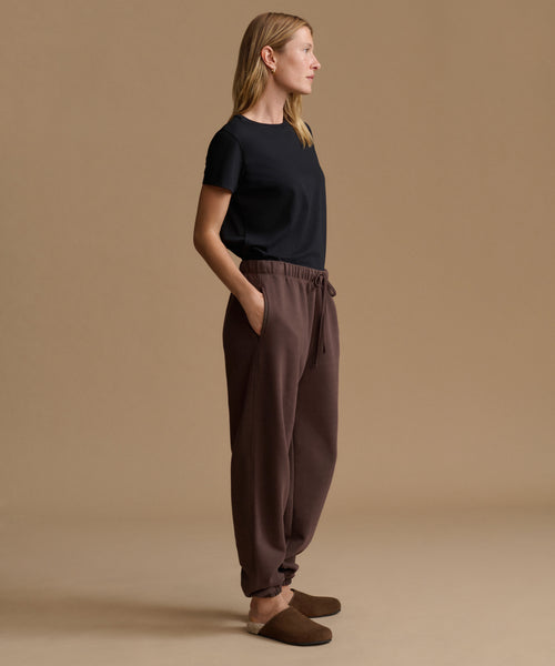 A woman stands in profile wearing a black short-sleeve top, brown drawstring jogger pants, and brown slip-on slippers against a plain beige studio background.