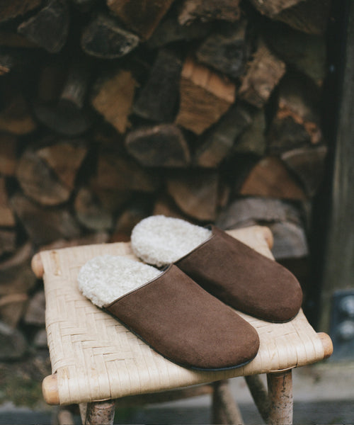 A pair of brown suede slip-on clogs with cream sherpa lining, displayed on a small woven stool in front of a stacked wood fire.