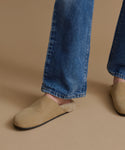 A close-up of a person wearing beige suede slip-on slippers with a soft fuzzy lining, paired with blue jeans, against a solid tan background.