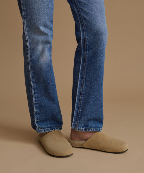 A person wearing blue denim jeans and beige suede slip-on clogs against a solid warm beige studio background.
