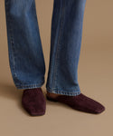 A person wearing blue jeans and dark burgundy suede mule flats, standing against a solid beige background.