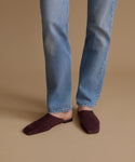 A person wearing light blue jeans and dark burgundy suede mule flats, standing against a solid beige studio background.