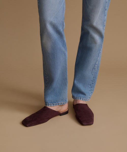 A person wearing light blue jeans and dark burgundy suede mule flats, standing against a solid beige studio background.