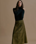 A woman modeling a black long-sleeve mock-neck top and an olive green A-line skirt against a solid beige studio background.