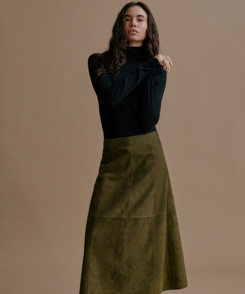 A woman modeling a black long-sleeve mock-neck top and an olive green A-line skirt against a solid beige studio background.