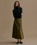 A woman stands against a plain warm beige studio background wearing a black long-sleeve mock-neck top and an olive green midi skirt with large patch pockets, paired with dark loafers.