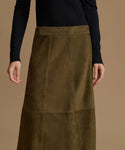 A model wearing a dark olive green suede midi skirt with a wide waistband and a black long-sleeve top, shown against a solid warm brown background.