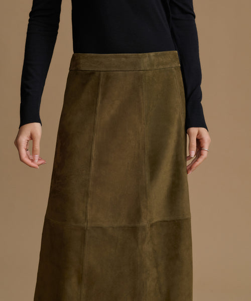 A model wearing a dark olive green suede midi skirt with a wide waistband and a black long-sleeve top, shown against a solid warm brown background.