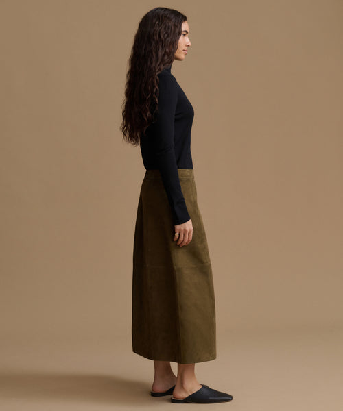 A woman stands in profile wearing a black long-sleeve top and a wide, knee-length olive green skirt against a plain beige studio background.