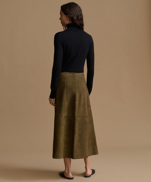 A woman shown from the back wearing a black long-sleeve turtleneck sweater and an olive green midi skirt, standing against a solid beige studio background.