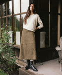 A woman stands on a porch wearing a cream knit sweater, a brown midi skirt, and black ankle boots, posed in front of a rustic house with large windows.