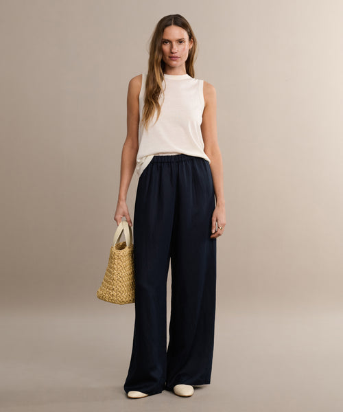 A woman poses casually in a sleeveless white top and navy, wide-leg Summer Demi Pant with a linen texture, paired with cream shoes and a woven straw bag, set against a neutral background. She has long hair.