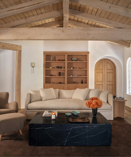 A styled living room featuring a cream-colored fabric sofa, a black marble coffee table, a beige armchair, and a large wooden bookshelf against a white wall.