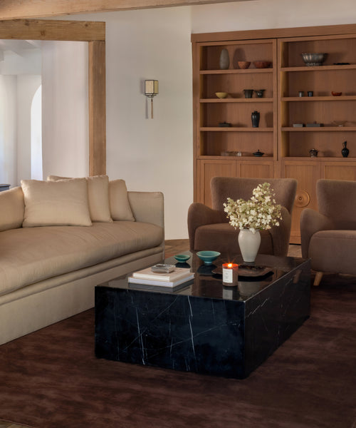A living room setup featuring a low rectangular black marble coffee table, a beige sofa, two brown armchairs, and a large wooden bookshelf in the background.