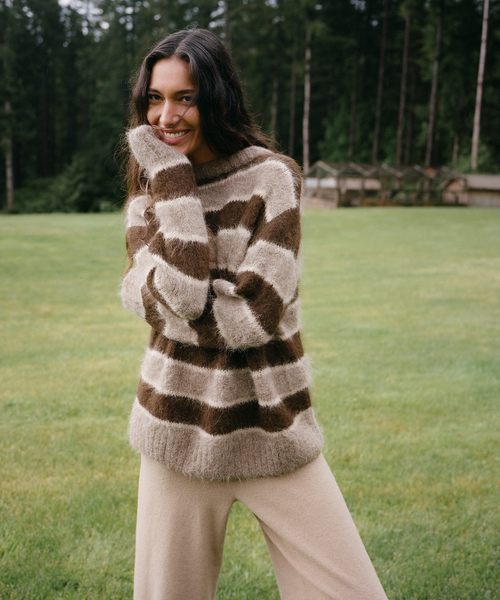A woman wearing a fuzzy brown and beige striped sweater and beige pants, standing in a grassy park with a forest in the background.