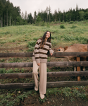 A woman stands outdoors on a grassy hill beside a wooden fence, wearing a brown and beige striped knit sweater and beige wide-leg pants. A cow is visible behind the fence, and a forested hill rises in the background.