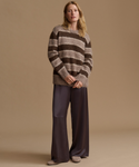 A woman stands against a warm beige studio background wearing a fuzzy striped knit sweater in brown and taupe tones, paired with dark wide-leg satin pants and taupe flats.