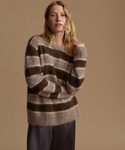 A woman wearing a fuzzy, oversized knit sweater with horizontal brown and beige stripes, paired with dark gray trousers, set against a solid warm beige studio background.