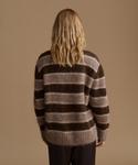 A woman viewed from the back wearing a fuzzy, long-sleeve knit sweater with horizontal brown and taupe stripes, set against a solid warm beige background.