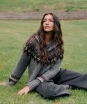 A woman sits on a grassy lawn wearing a gray knit sweater with a brown and cream Fair Isle pattern around the yoke and cuffs, paired with dark gray wide-leg pants.