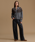 A model stands against a solid tan studio background wearing a gray knit cardigan with a brown and white Fair Isle yoke pattern, paired with black wide-leg trousers and brown suede shoes.