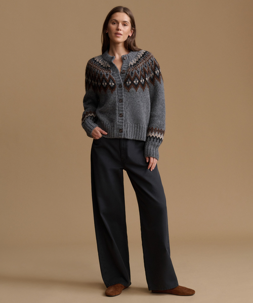 A model stands against a solid tan studio background wearing a gray knit cardigan with a brown and white Fair Isle yoke pattern, paired with black wide-leg trousers and brown suede shoes.