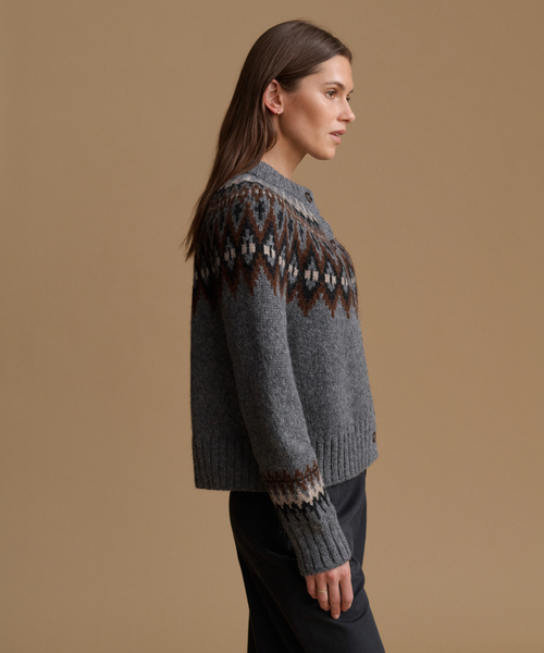 A woman in side profile wearing a gray knit cardigan with a brown and cream geometric Fair Isle pattern across the shoulders and cuffs, set against a solid tan studio background.