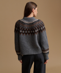 A woman shown from the back wearing a gray knit sweater with a brown and cream Fair Isle yoke pattern, paired with black trousers, against a solid beige studio background.