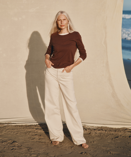 Model wearing a brown long-sleeve t-shirt with cream color jeans at the beach.
