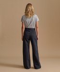 A woman viewed from behind wearing a light gray short-sleeve t-shirt and dark charcoal wide-leg jeans, standing against a solid beige studio background.