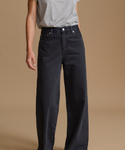 A model wearing high-waisted, wide-leg black denim jeans with a gray t-shirt, shown from the waist down against a solid beige studio background.