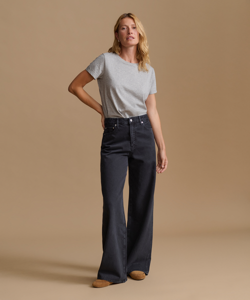 A woman modeling a pair of dark charcoal wide-leg jeans with a light gray t-shirt, standing against a solid beige studio background.