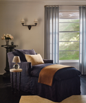 A dark navy slipcovered chaise lounge chair in a styled room, featuring a beige throw pillow, a tan blanket draped over the foot, a small side table with a lamp, and a window with sheer curtains.