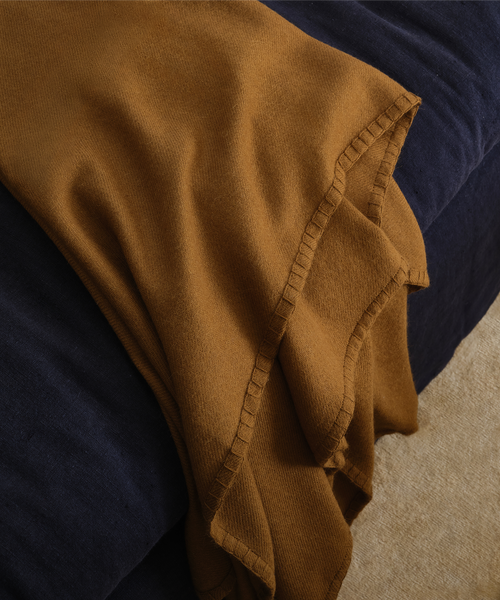 A close-up of a warm camel brown knit throw blanket draped over a dark navy blue chaise, showing its soft texture and decorative stitched border.