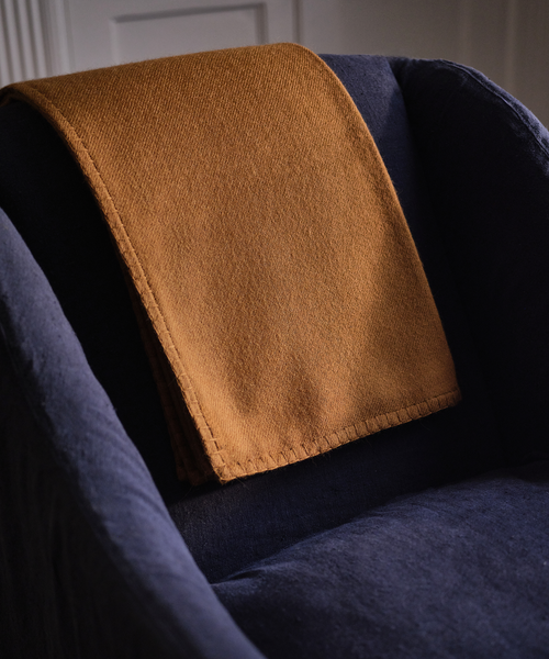 A folded mustard yellow wool blanket with visible stitching rests on a dark navy blue armchair in a dimly lit interior setting.