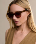 A woman with long blonde hair wears oversized Thompson Sunglasses featuring UV protection, a sleeveless tan top, and small hoop earrings as she poses against a plain beige background.